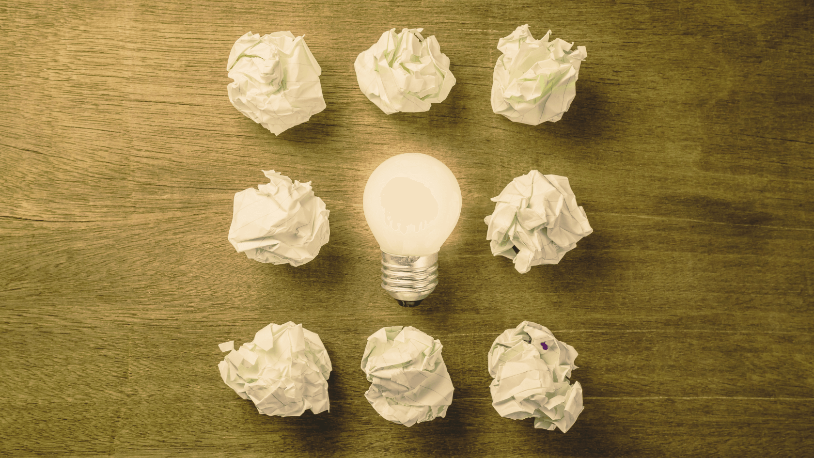 Crumpled paper balls surrounding a glowing light bulb on a wooden surface.